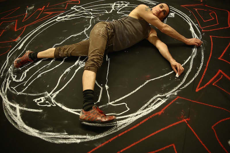 John Kelly lays on the ground and draws around himself with red and white chalk. Various lines and designs flank him.